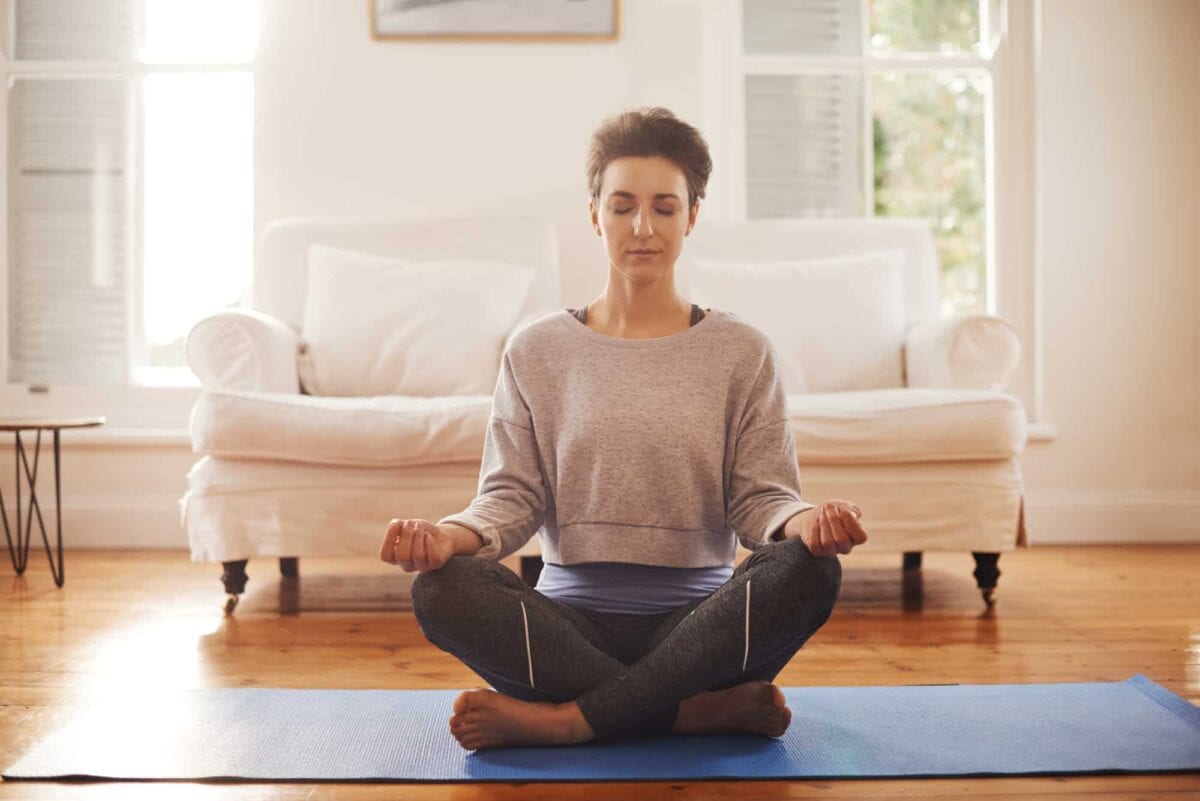 A person sits cross-legged on a yoga mat in a living room with eyes closed, meditating in front of a white sofa and windows.