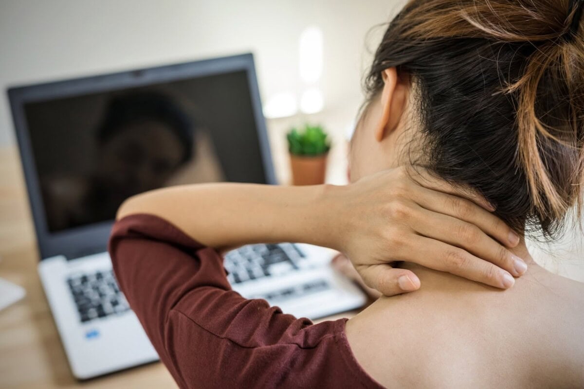 A woman sits at a desk in front of a laptop, holding the back of her neck as if experiencing discomfort or pain.
