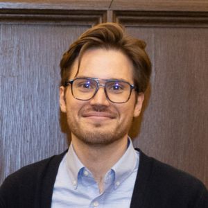 A man with glasses, light brown hair, and a short beard is standing in front of a wooden wall, wearing a light blue shirt and dark sweater.