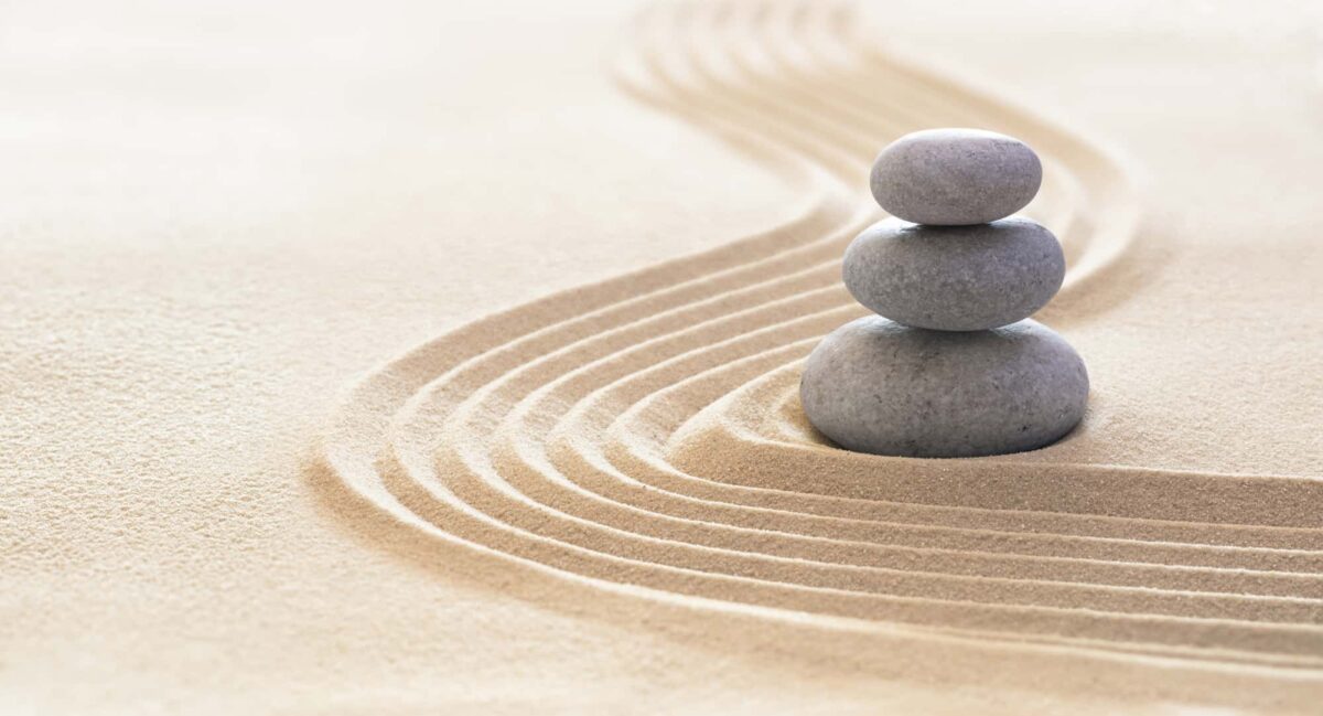 Three smooth stones stacked on raked sand with parallel lines curving around them, creating a minimalist and serene composition.